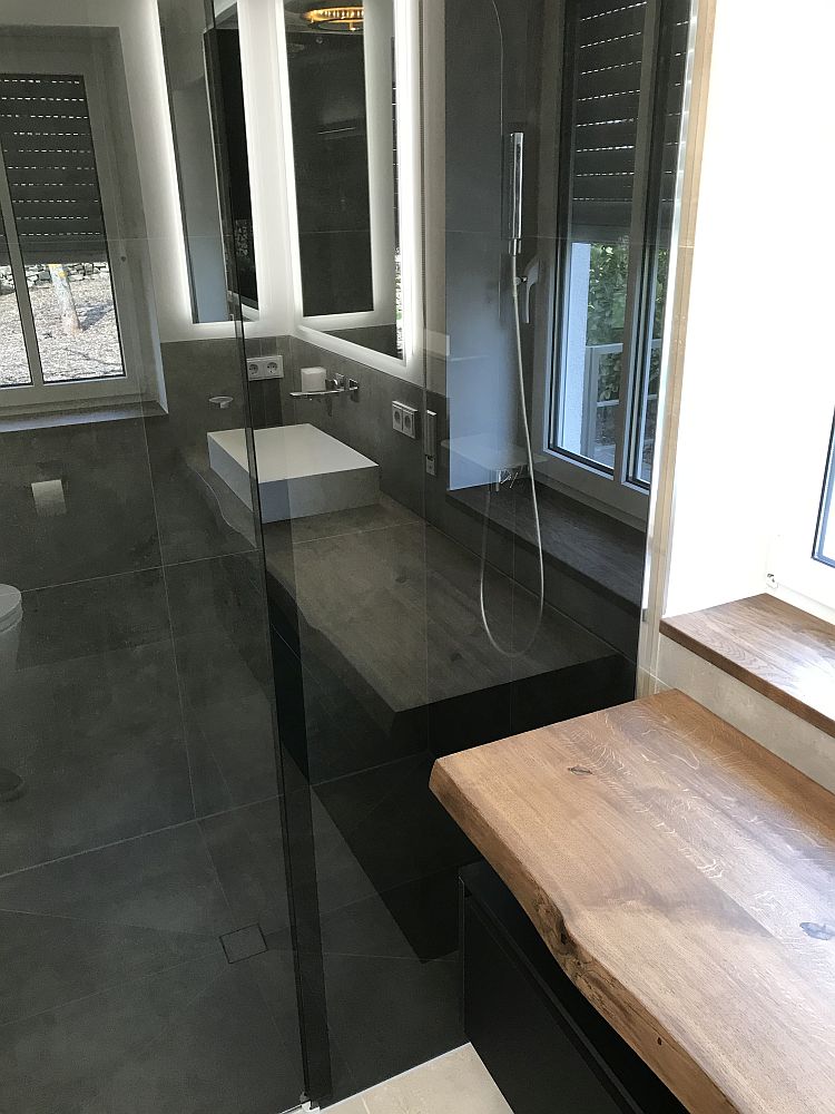 Badezimmer mit bodengleicher Dusche, Glasabtrennung, natürlichem Holz-Waschtisch, schwarzen Armaturen und großen Fenstern für viel Tageslicht.