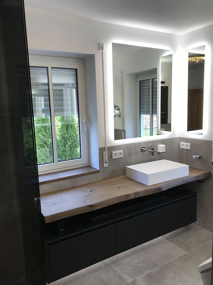 Modernes Masterbad mit einem massiven Holz-Waschtisch, einem weißen rechteckigen Waschbecken, beleuchteten Spiegeln und einem großen Fenster mit Blick ins Grüne.