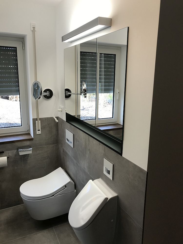 Minimalistisches Badezimmer mit einer Spiegelwand, einem wandhängenden WC und Bidet, sowie natürlichem Licht, das durch ein Fenster mit Jalousien fällt.