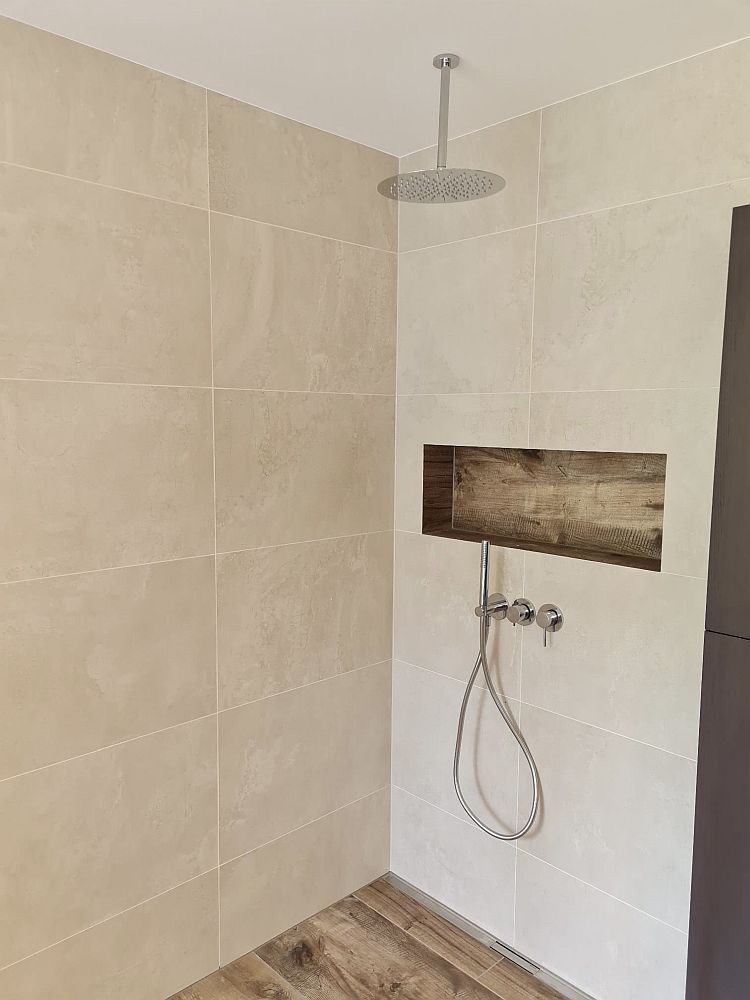 Eine moderne Dusche mit einer Regendusche, die von der Decke herabhängt, und einer Wand aus großformatigen, beigen Fliesen.