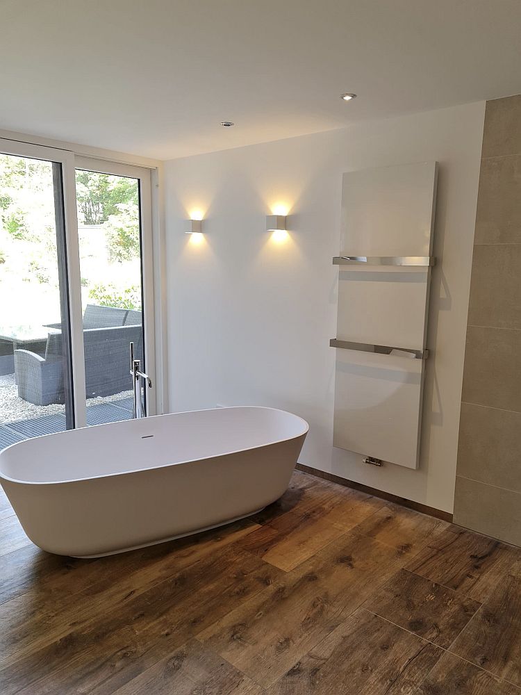 Badezimmer mit freistehender Wanne, Parkettboden und Glas-Schiebetür. Wandlampen und Handtuchheizkörper sorgen für eine stilvolle Atmosphäre.