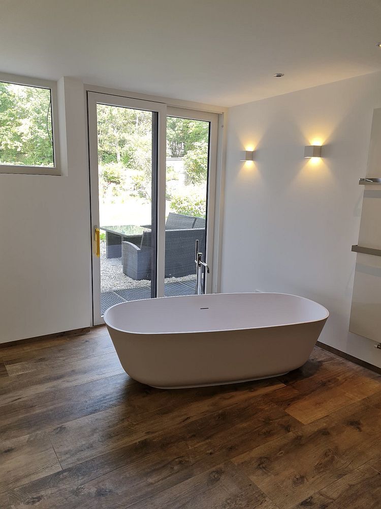 Eine luxuriöse Badewanne steht frei im Raum vor einer großen Glas-Schiebetür, die auf eine Terrasse mit Gartenblick führt.