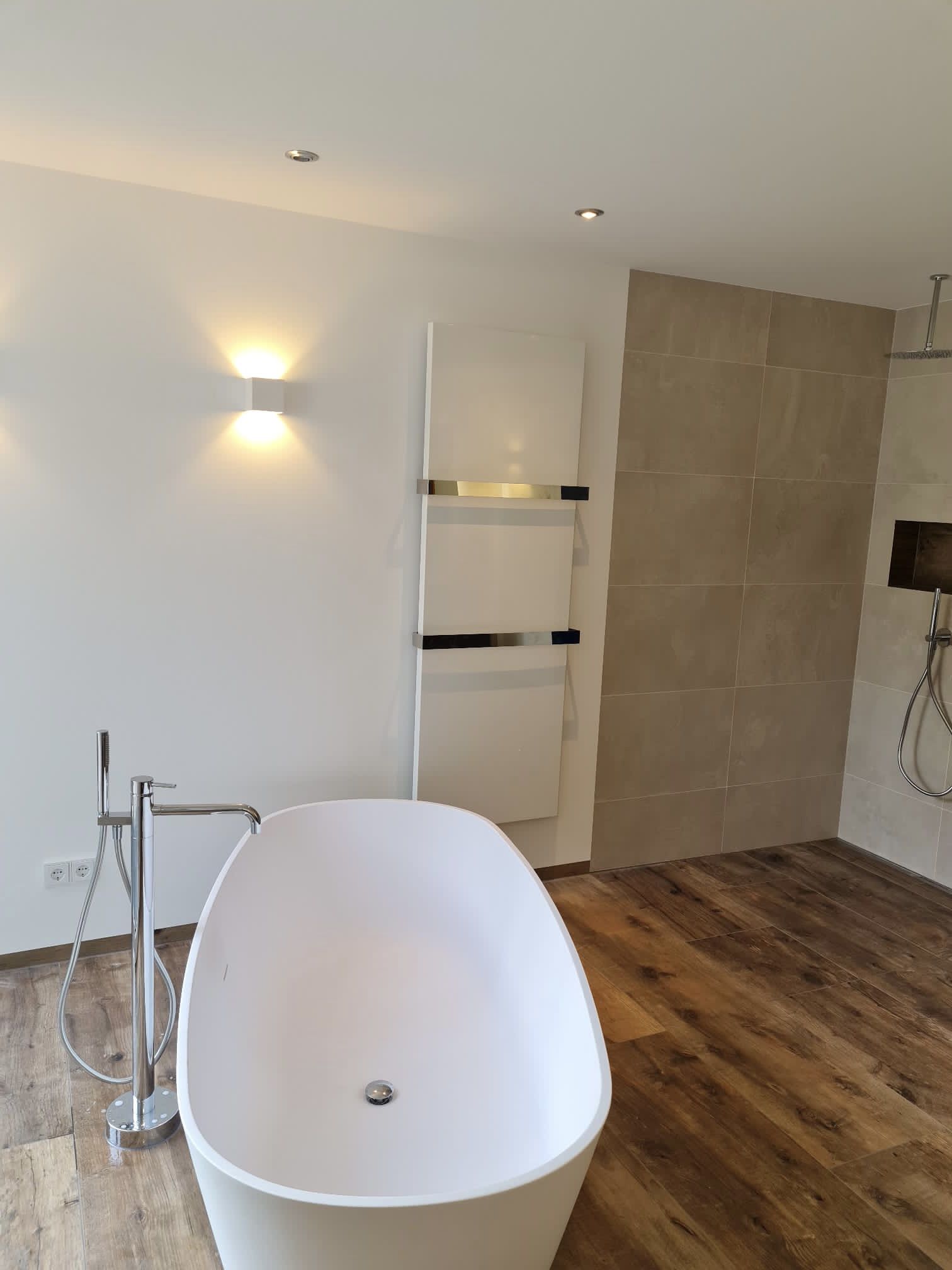 Badewanne mit einer freistehenden Armatur aus Edelstahl. Daneben befindet sich eine bodenebene Walk-in-Dusche.