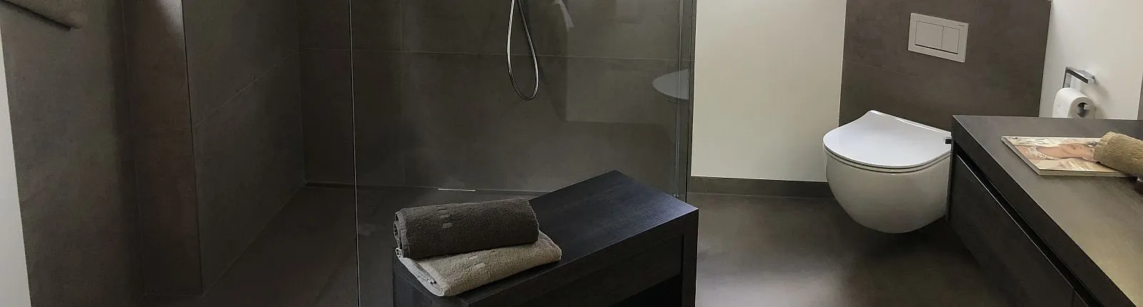 Elegante Walk-In-Dusche mit Glasabtrennung, Wand-WC und dunklem Waschtisch. Eine Sitzbank mit Handtüchern ergänzt das stilvolle Design in Grautönen.