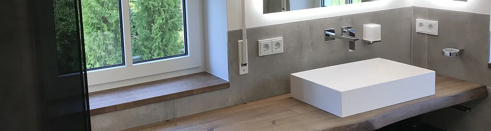 Ein modernes Masterbad mit Betonoptik-Wand, schwebendem Holz-Waschtisch, einem eckigen weißen Waschbecken und einem großen Fenster mit Blick ins Grüne.