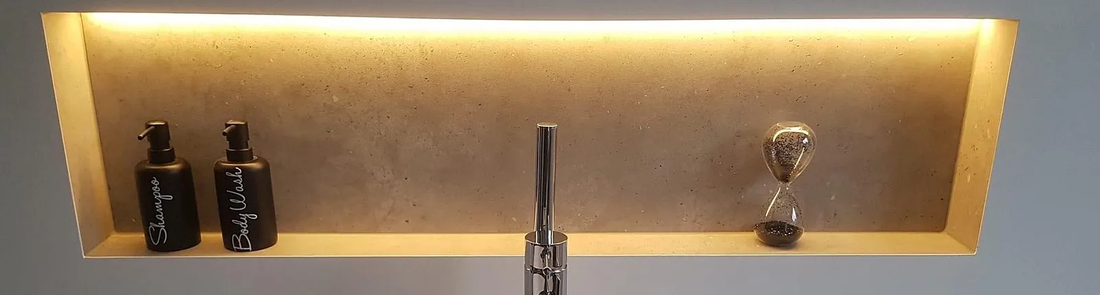 Beleuchtete Wandnische mit warmem LED-Licht, in der eine Sanduhr und zwei schwarze Spenderflaschen für Shampoo und Body Wash stehen.