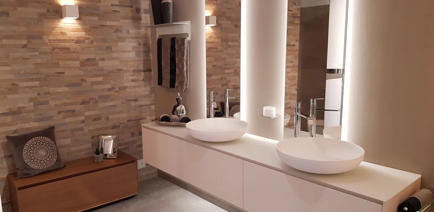 Das Bild zeigt ein Badezimmer mit einem weißen Waschtisch, runden Waschbecken, hinterleuchteten Spiegeln, einer Natursteinwand, Holzbank und dezenter Beleuchtung.