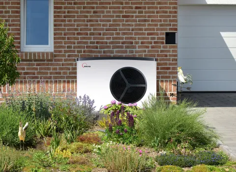 Eine moderne Wärmepumpe steht neben einem gepflegten Blumenbeet vor einem Ziegelhaus mit weißer Garage.