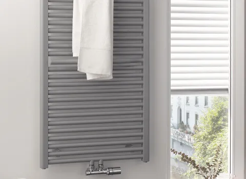 In einem hellen Badezimmer hängt ein grauer HSK Line Plus Heizkörper mit einem weißen Handtuch daran. 