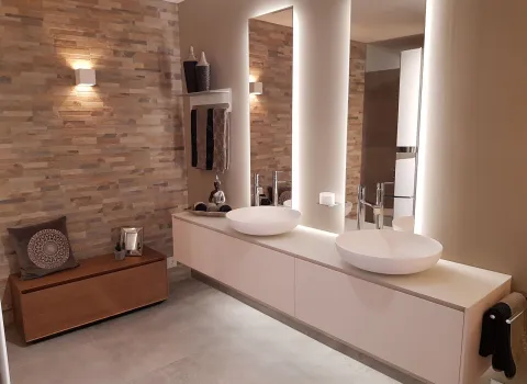 Das Bild zeigt ein Badezimmer mit einem weißen Waschtisch, runden Waschbecken, hinterleuchteten Spiegeln, einer Natursteinwand, Holzbank und dezenter Beleuchtung.