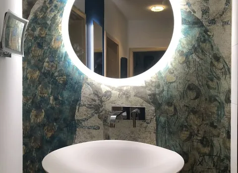 Ein stilvoller Waschtisch mit rundem, beleuchtetem Spiegel vor einer Wandgestaltung mit Pfauenmotiv.