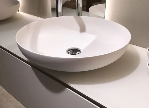 Ein elegantes, rundes Aufsatzwaschbecken aus weißem Material mit modernen, verchromten Armaturen auf einem minimalistischen Waschtisch.