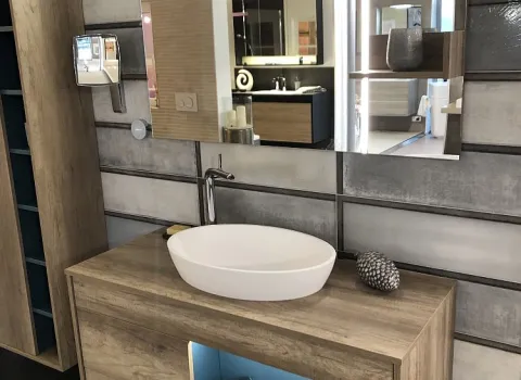 Ein moderner Waschtisch in Holzoptik mit offenem Fach, beleuchtetem Spiegel und minimalistischen Accessoires vor einer strukturierten Wandgestaltung.