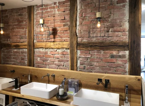 Ein rustikaler Doppelwaschtisch aus Holz vor einer Wand aus rotem Backstein mit sichtbaren Holzbalken, ergänzt durch Käfiglampen im Industrial-Stil.