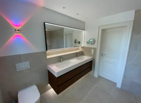 Seitliche Perspektive auf einen massiven Doppelwaschtisch, einem großem Spiegel, wandhängendem WC und halbhohen hell-beigen Fliesen.