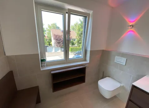 Vom Sanitär- und Heizungshandwerksunternehmen smr geplantes und realisiertes Badezimmer in Schweinfurt mit Holzmöbeln, rosa Licht, wandhängendem WC und halbhohen hell-beigen Fliesen.