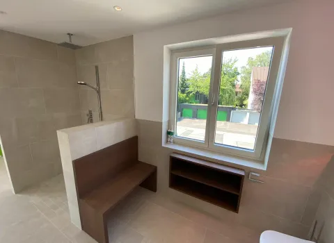 Vom Sanitär- und Heizungshandwerksunternehmen smr geplantes und realisiertes Badezimmer in Schweinfurt mit Sitzbank und Regal aus Holz, Regendusche, wandhängendem WC und hell-beigen Fliesen.