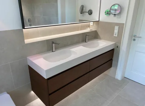 Seitliche Perspektive auf einen massiven Doppelwaschtisch und einen großen Spiegel vor halbhohen hell-beigen Fliesen.