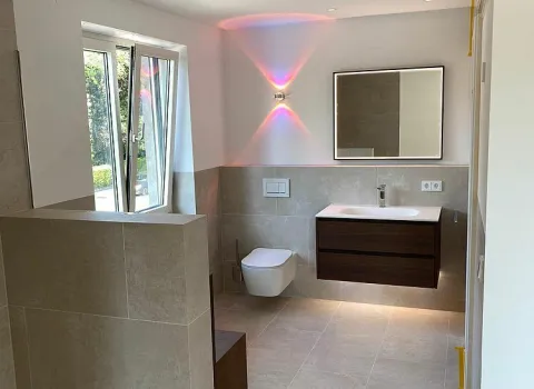 Vom Sanitär- und Heizungshandwerksunternehmen smr GmbH geplantes und realisiertes Masterbadezimmer in Schweinfurt mit massivem Waschtisch, großem Spiegel, wandhängendem WC und halbhohen hell-beigen Fliesen.