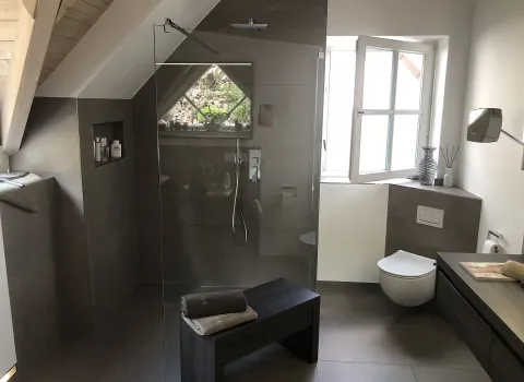 Dachgeschoss-Badezimmer mit begehbarer Dusche, Wand-WC und dunkler Sitzbank. Graue Fliesen und Holzdetails verleihen dem Raum eine warme Eleganz.