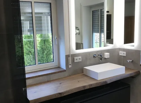 Modernes Masterbad mit einem massiven Holz-Waschtisch, einem weißen rechteckigen Waschbecken, beleuchteten Spiegeln und einem großen Fenster mit Blick ins Grüne.