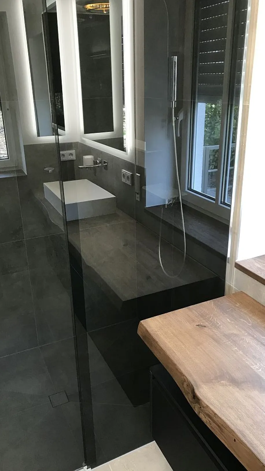 Badezimmer mit bodengleicher Dusche, Glasabtrennung, natürlichem Holz-Waschtisch, schwarzen Armaturen und großen Fenstern für viel Tageslicht.
