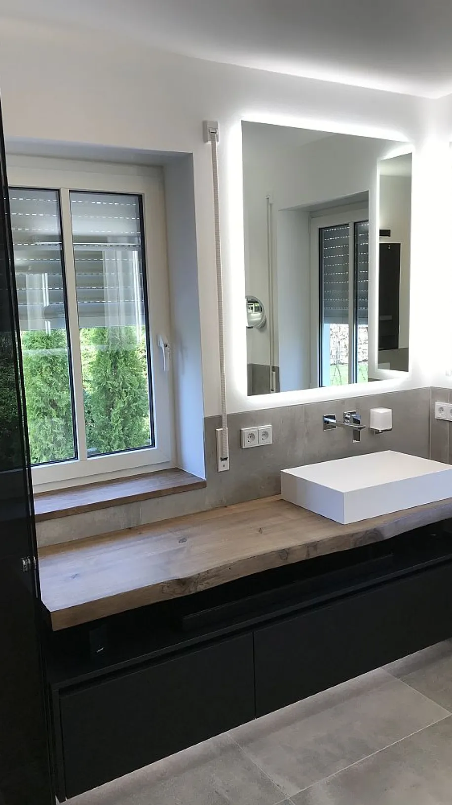 Modernes Masterbad mit einem massiven Holz-Waschtisch, einem weißen rechteckigen Waschbecken, beleuchteten Spiegeln und einem großen Fenster mit Blick ins Grüne.