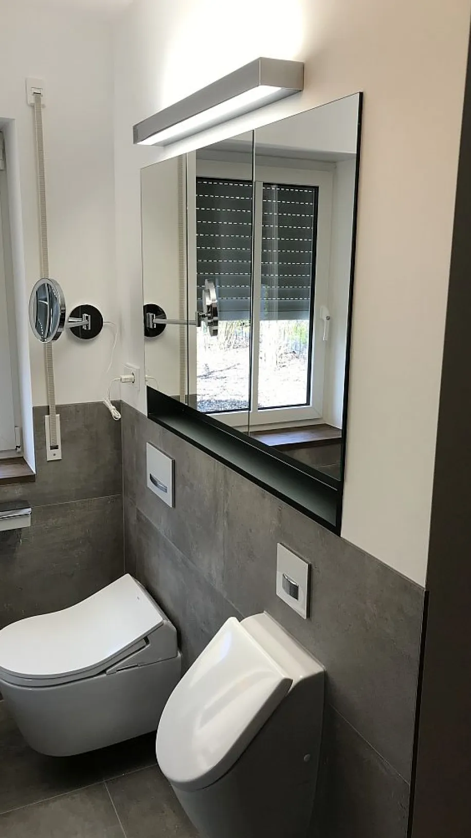 Minimalistisches Badezimmer mit einer Spiegelwand, einem wandhängenden WC und Bidet, sowie natürlichem Licht, das durch ein Fenster mit Jalousien fällt.