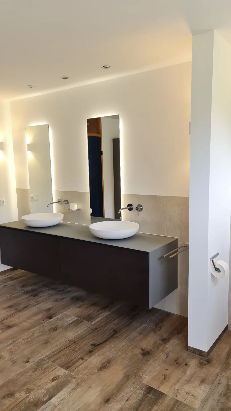 Waschplatz mit wandhängendem Doppelwaschtisch, LED-beleuchteten Spiegeln und Parkettboden neben einer Trennwand.