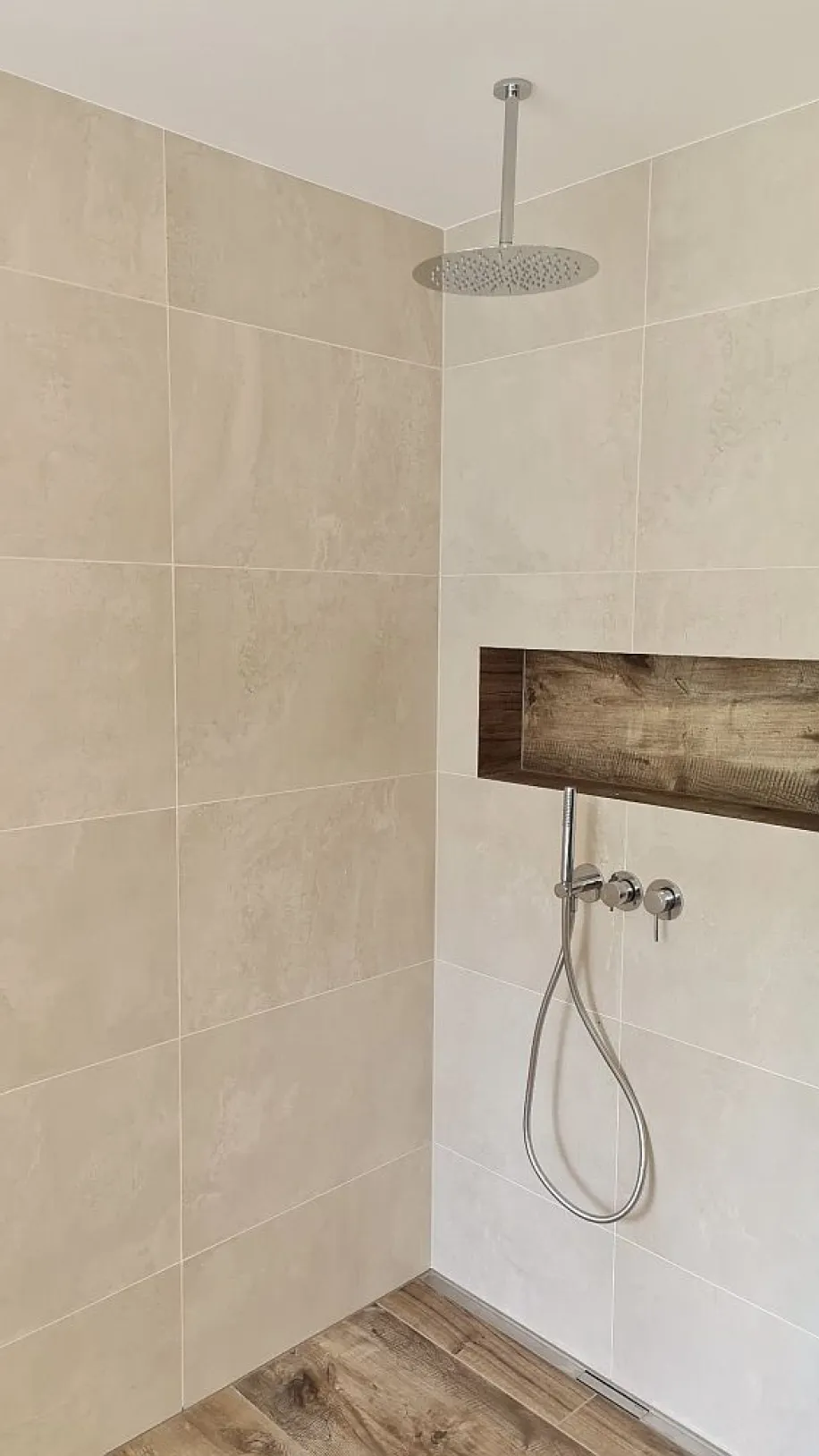 Eine moderne Dusche mit einer Regendusche, die von der Decke herabhängt, und einer Wand aus großformatigen, beigen Fliesen.