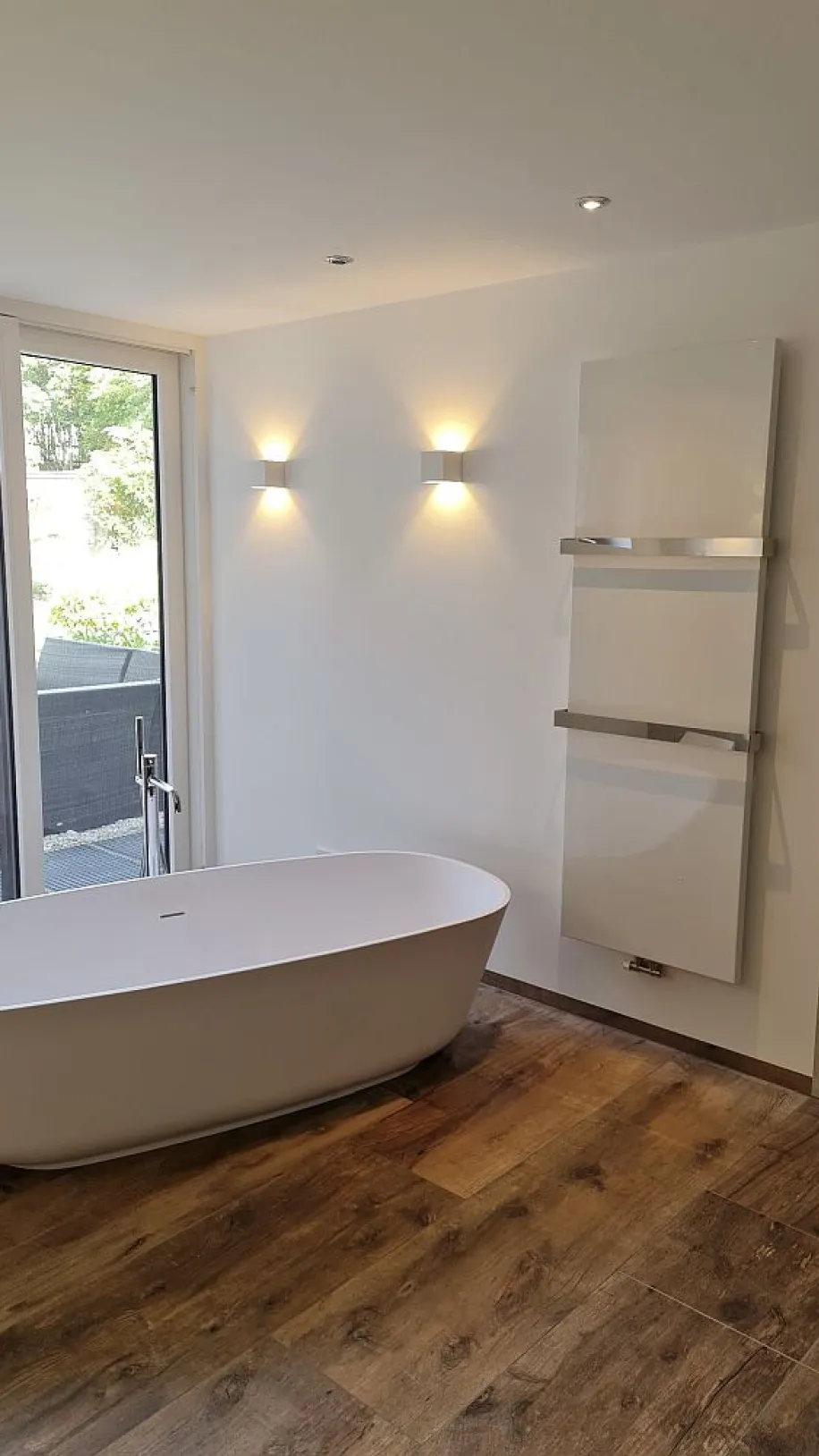 Badezimmer mit freistehender Wanne, Parkettboden und Glas-Schiebetür. Wandlampen und Handtuchheizkörper sorgen für eine stilvolle Atmosphäre.