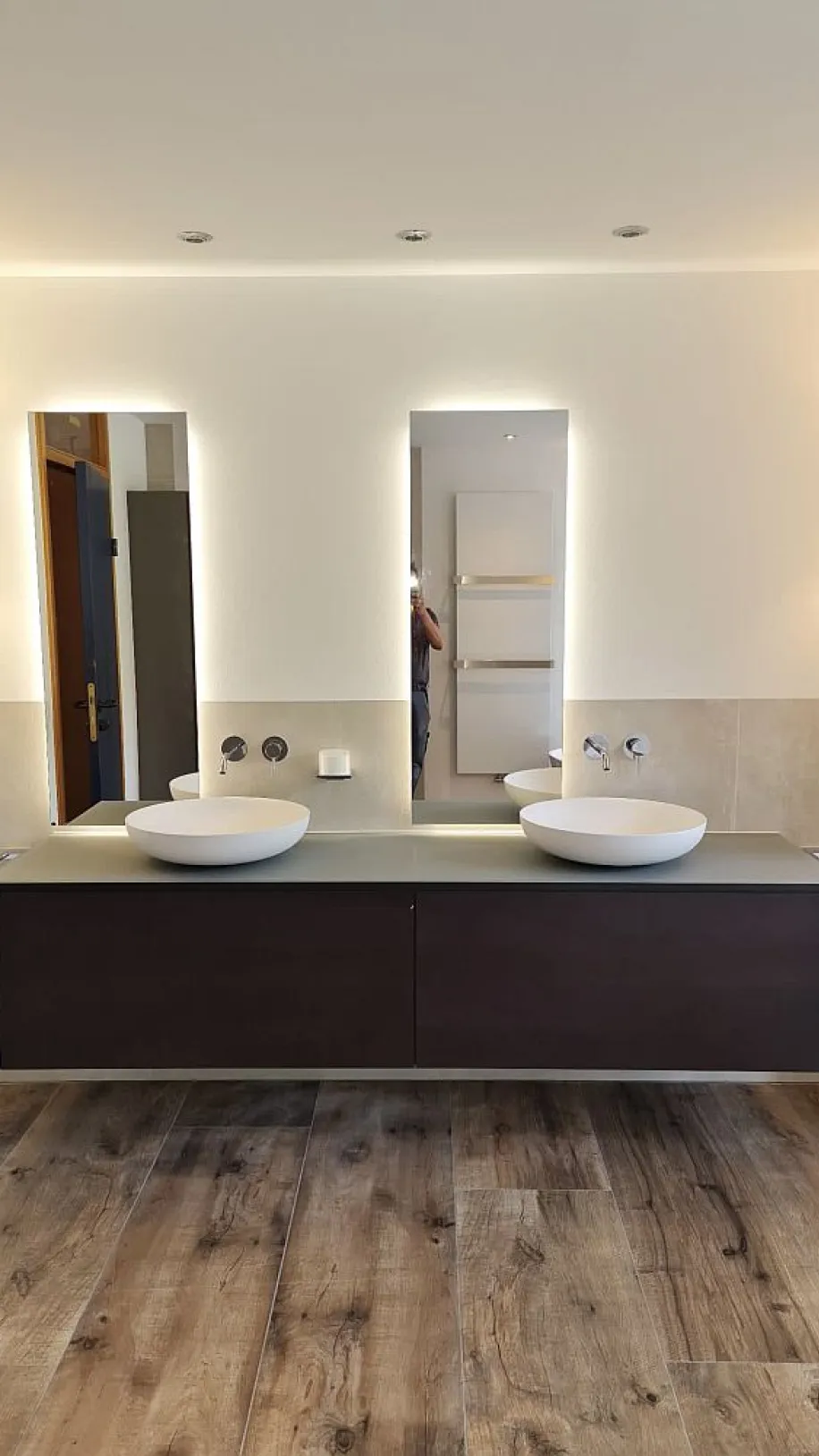 Ein elegantes Badezimmer mit einem wandmontierten Waschtisch in dunkler Holzoptik, darauf zwei stilvolle, runde Keramikwaschbecken. Der Boden ist mit hochwertigem Parkett ausgelegt.