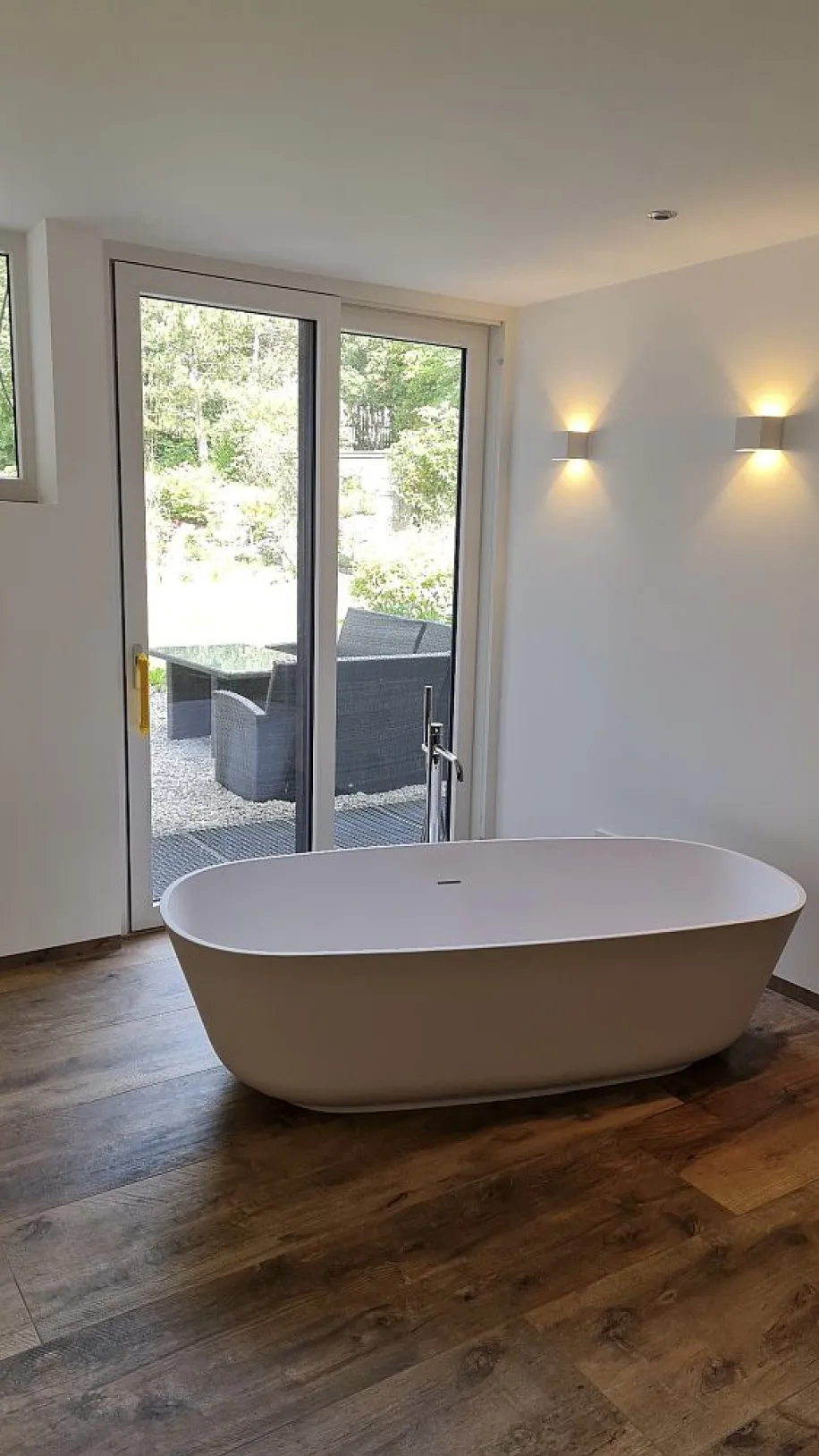 Eine luxuriöse Badewanne steht frei im Raum vor einer großen Glas-Schiebetür, die auf eine Terrasse mit Gartenblick führt.