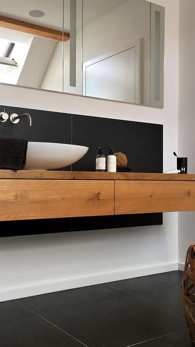 Helles Badezimmer mit einem minimalistischen Holz-Waschtisch, einem weißen Waschbecken, einer schwarzen Akzentwand und einem geflochtenen Sitzhocker.