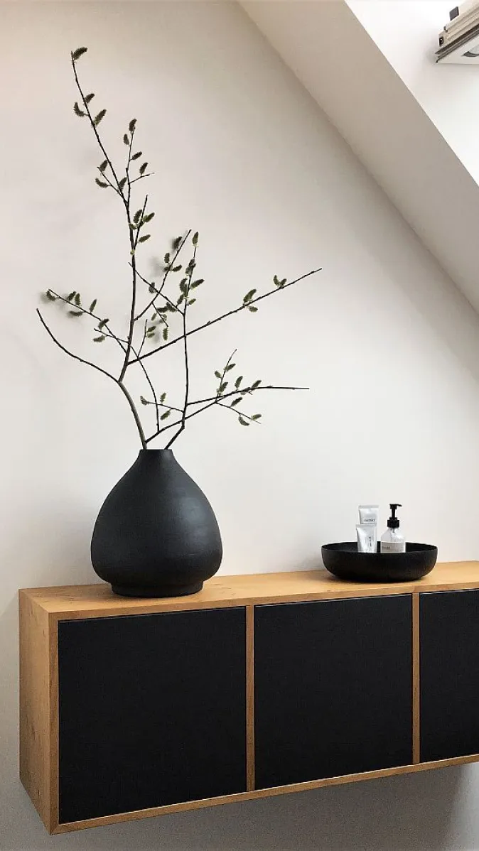 Minimalistisches Interieur mit einem hängenden Holzschrank, schwarzer Front, dekorativer schwarzer Vase mit Zweigen und natürlichem Licht durch ein Dachfenster.