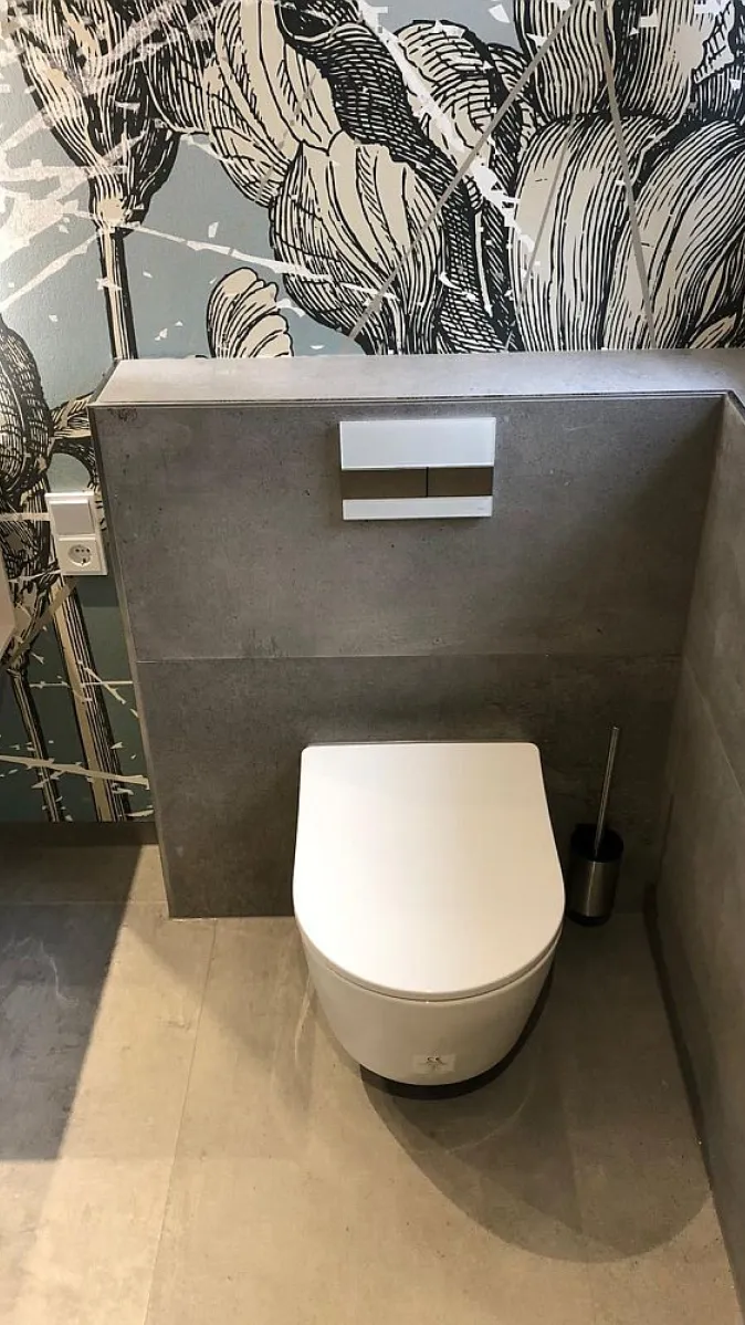 Die kompakte, minimalistische Toilette ist in einem Badezimmer mit grauen Fliesen und einer dekorativen Wand mit floralen Mustern untergebracht. 