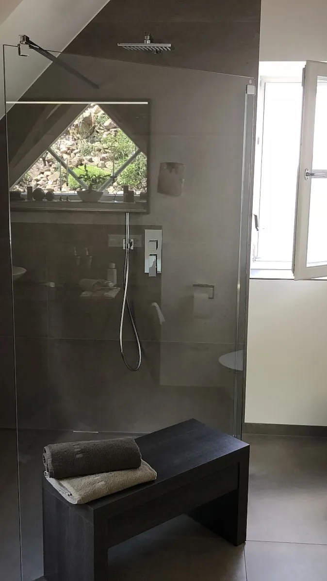 Dachgeschoss-Badezimmer mit begehbarer Dusche, Wand-WC und dunkler Sitzbank. Graue Fliesen und Holzdetails verleihen dem Raum eine warme Eleganz.