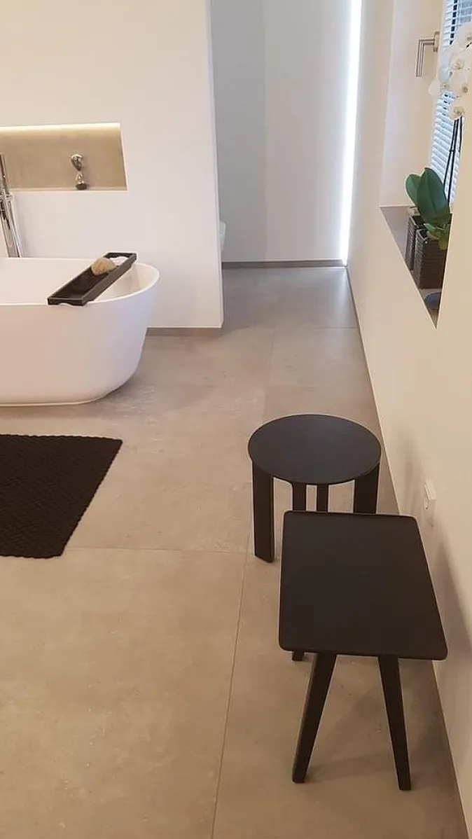 Minimalistisches Badezimmer mit großer Badewanne, beigen Fliesen, schwarzen Hockern und dekorativer Wandnische.