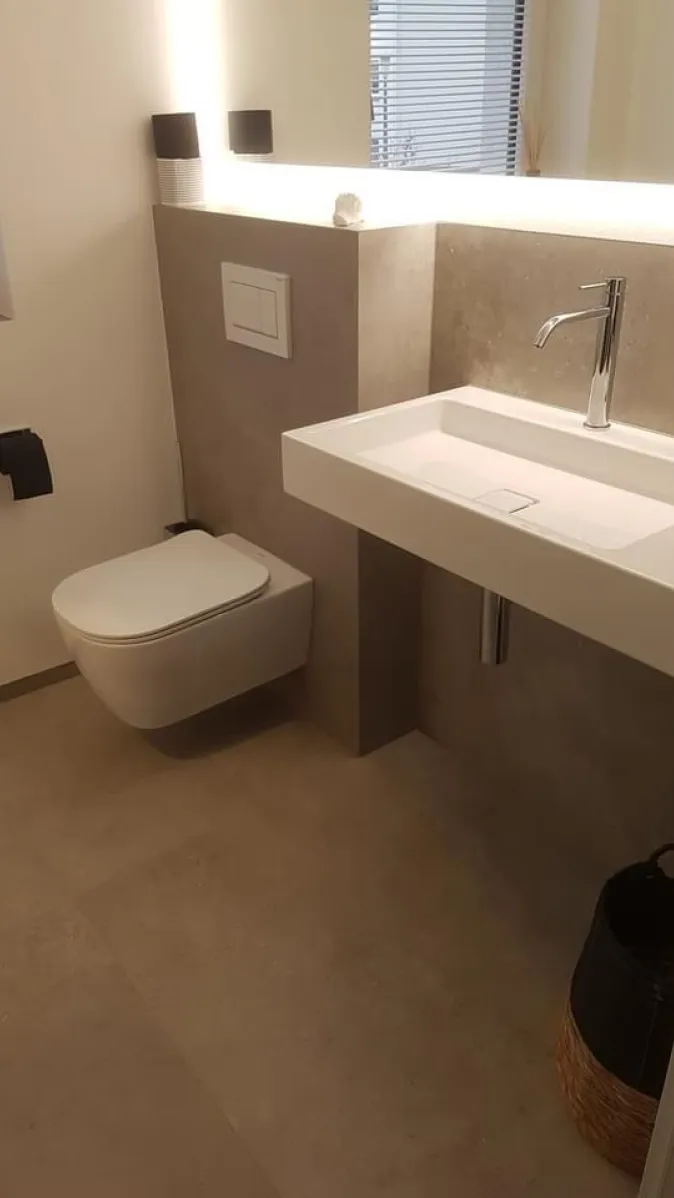 Elegantes WC mit Wandmontage, rechteckigem Waschtisch, beleuchtetem Spiegel und Fliesen in Beton-Optik.