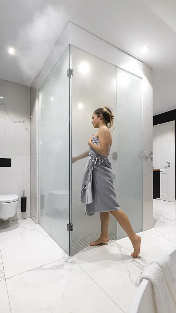 Eine Frau steht seitlich zu einer dampfbefüllten Dampfdusche und öffnet die Tür.