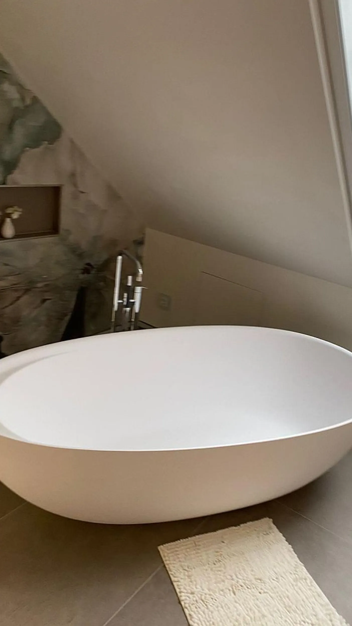 Die freistehende, weiße Badewanne in einem Dachbadezimmer steht auf großformatigen Fliesen vor einer stilvollen Wand mit natürlichen Grün- und Beigetönen.