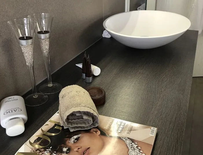 Stilvoll eingerichteter Waschtisch mit Glasdeko, einer gerollten Handtuchrolle und einer Zeitschrift.