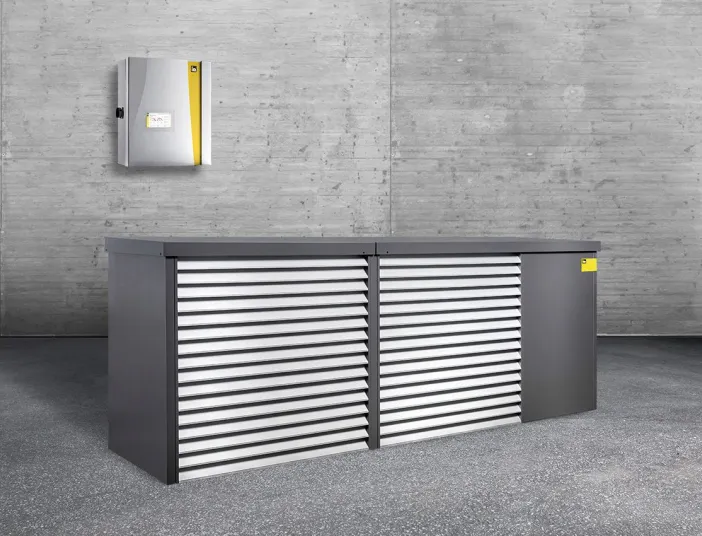 Eine Kaskadenlösung der intelligenten Luftwärmepumpe iDM Aero Alm Max steht in einem grauen Raum. Dahinter hängt die kleine Inneneinheit.